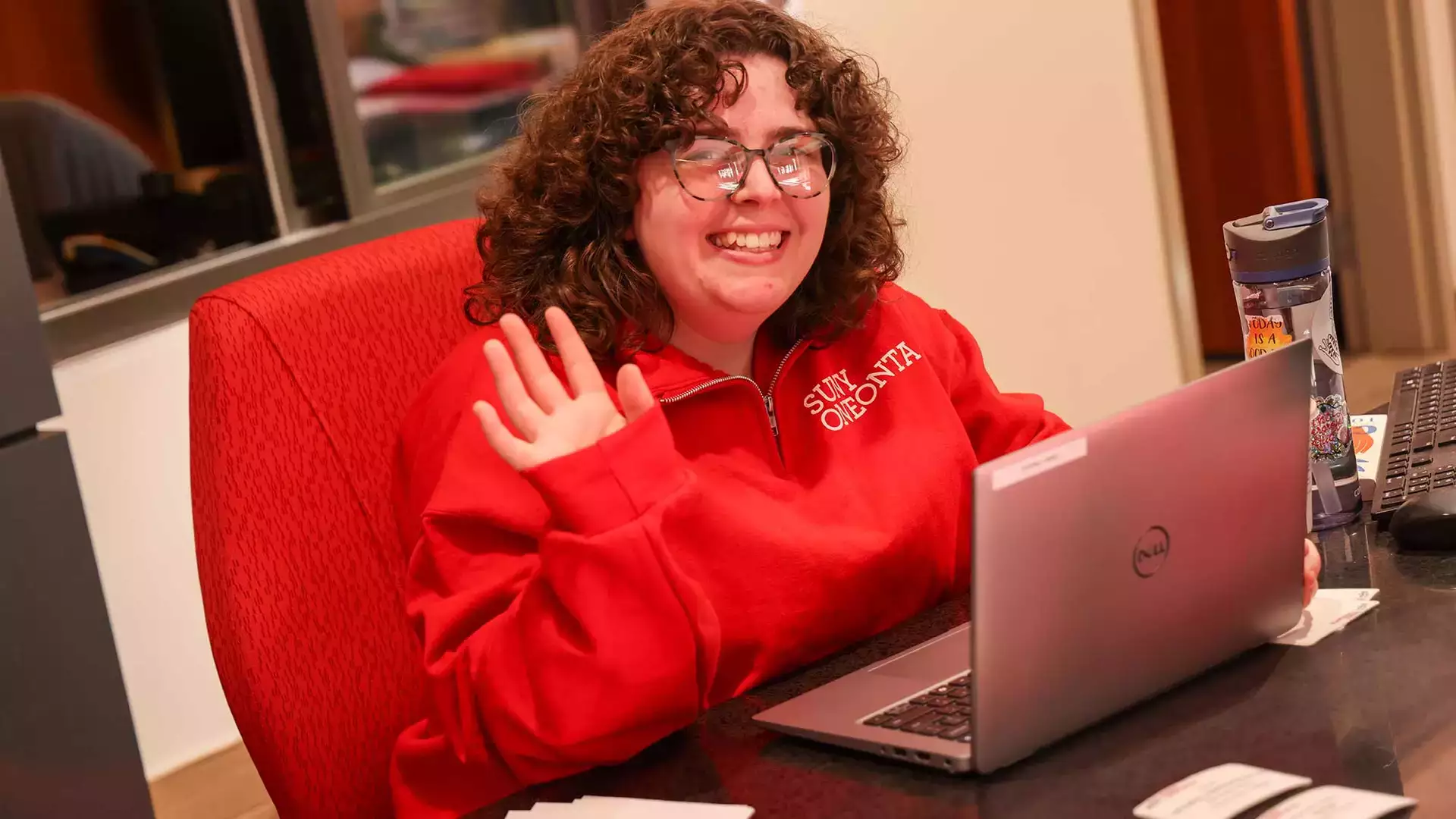 Person sitting at a desk smiling and waving "hi"