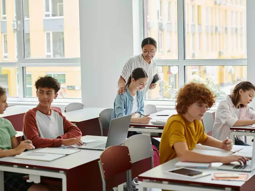 Smiling teacher standing next to adolescent students during lesson with devices in class