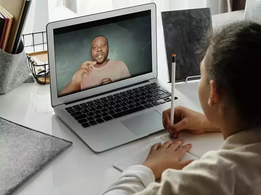 A child takes notes while watching their teacher on a laptop
